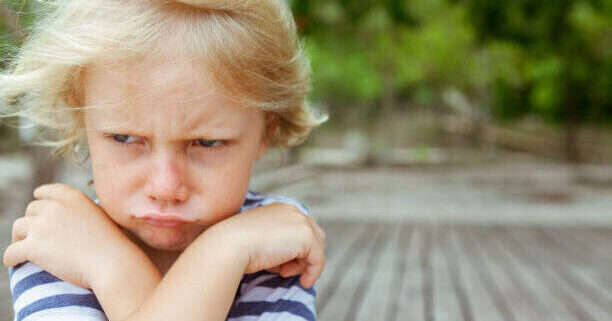 Face portrait of annoyed and unhappy caucasian kid with crossed arms. Upset and angry child concept for family relations, social problems issues and juvenile psychology.  Copy space outdoor background.